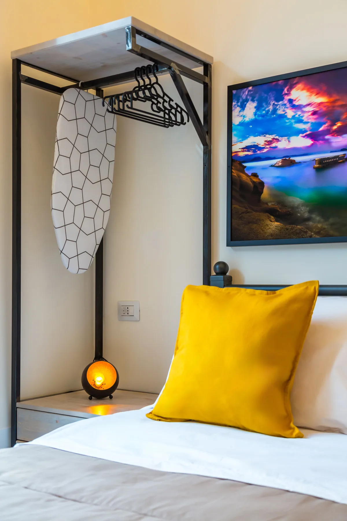 Un dormitorio con ropa de cama blanca, un cojín amarillo y un perchero de pared.