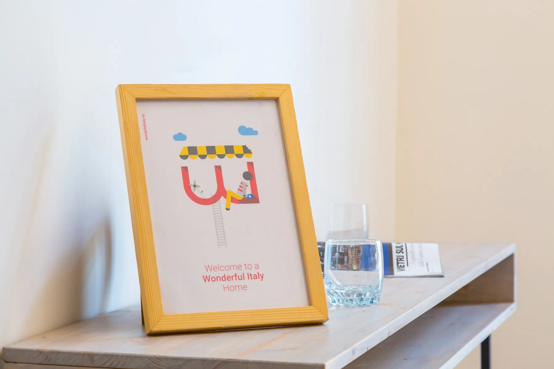 Una lámina ilustrada enmarcada y un vaso están sobre una repisa clara.