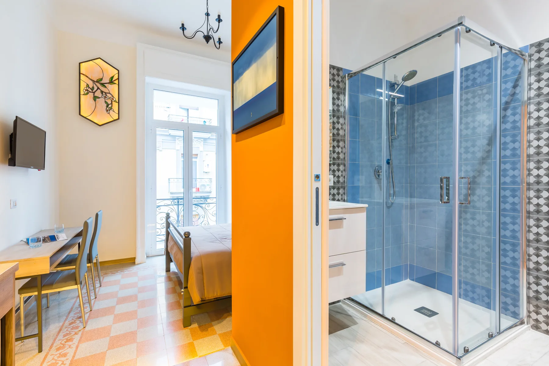 Una habitación luminosa con escritorio y cama da a una ducha acristalada con azulejos.