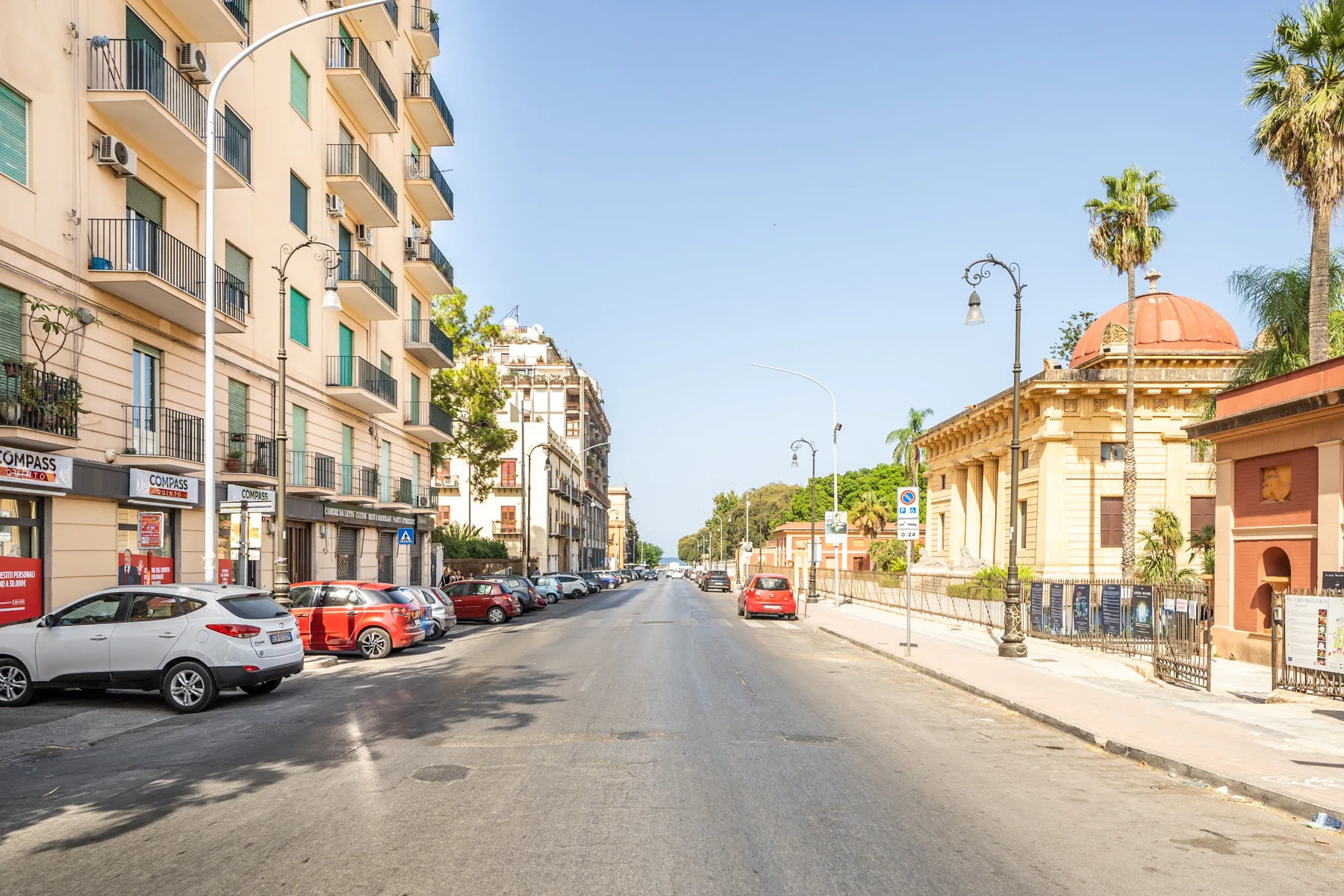 Una calle urbana flanqueada por edificios, coches estacionados y palmeras.