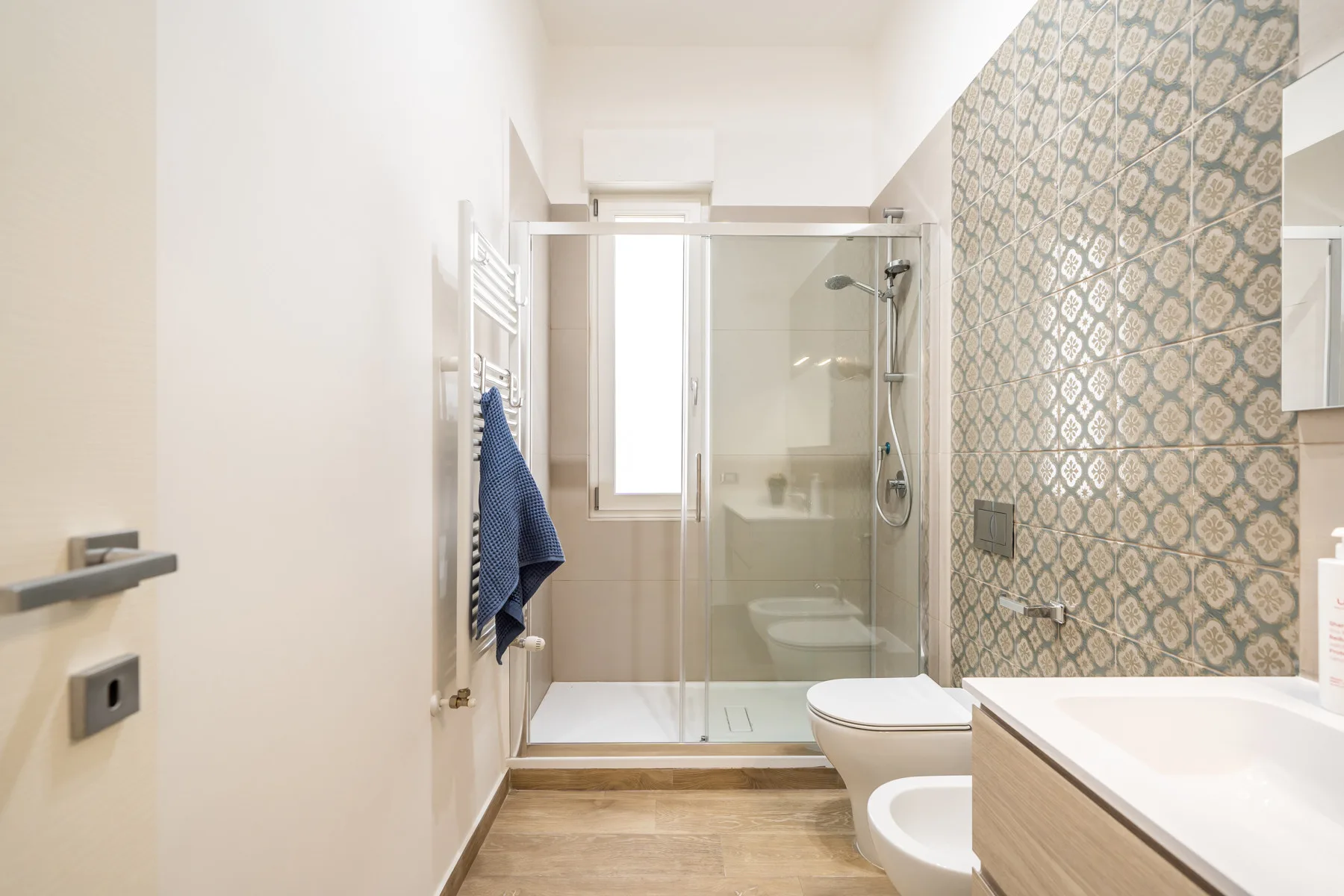 Un baño luminoso con ducha de vidrio y una pared de azulejos estampados.