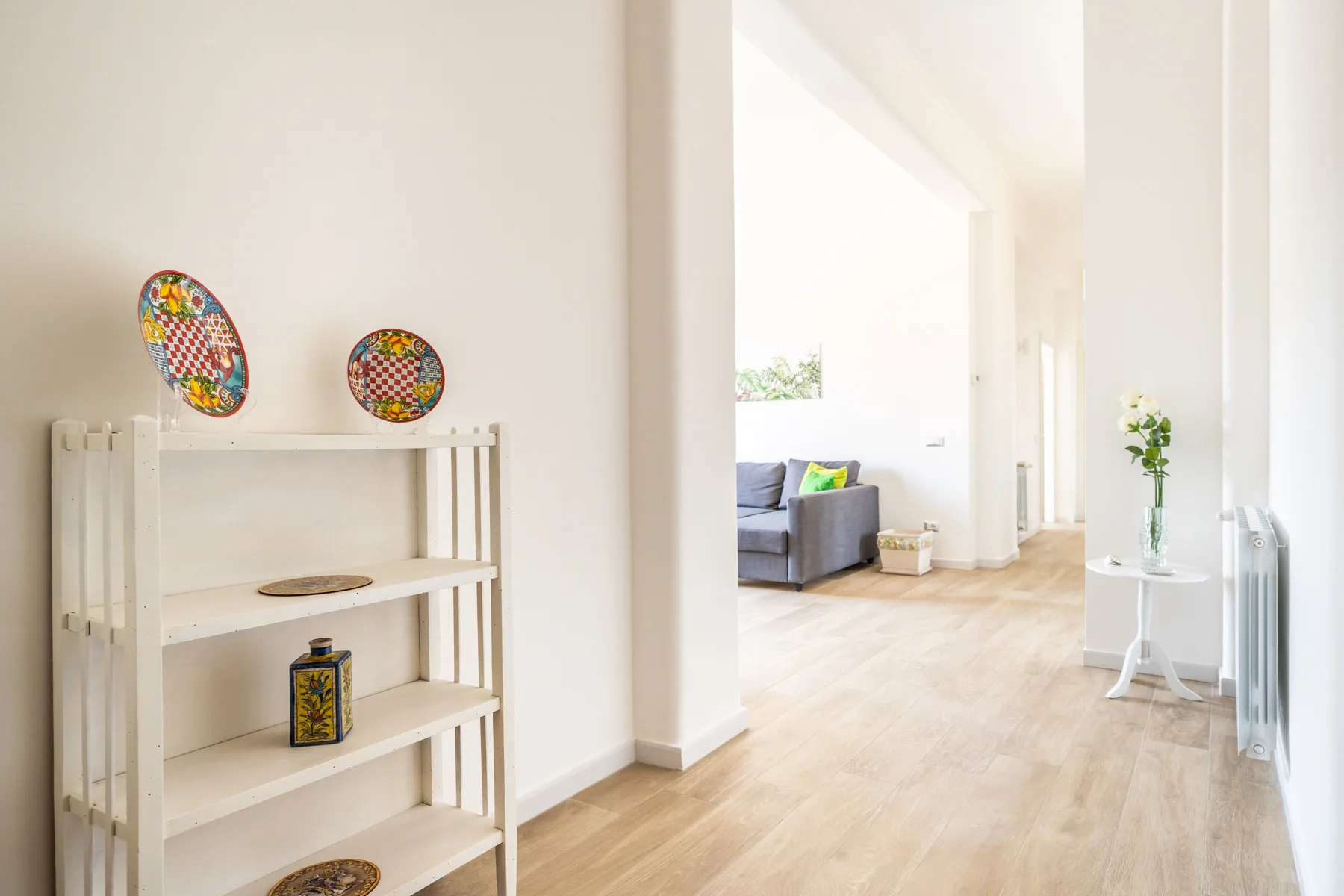 Un pasillo luminoso se abre a una sala de estar, con una estantería blanca junto a la pared.