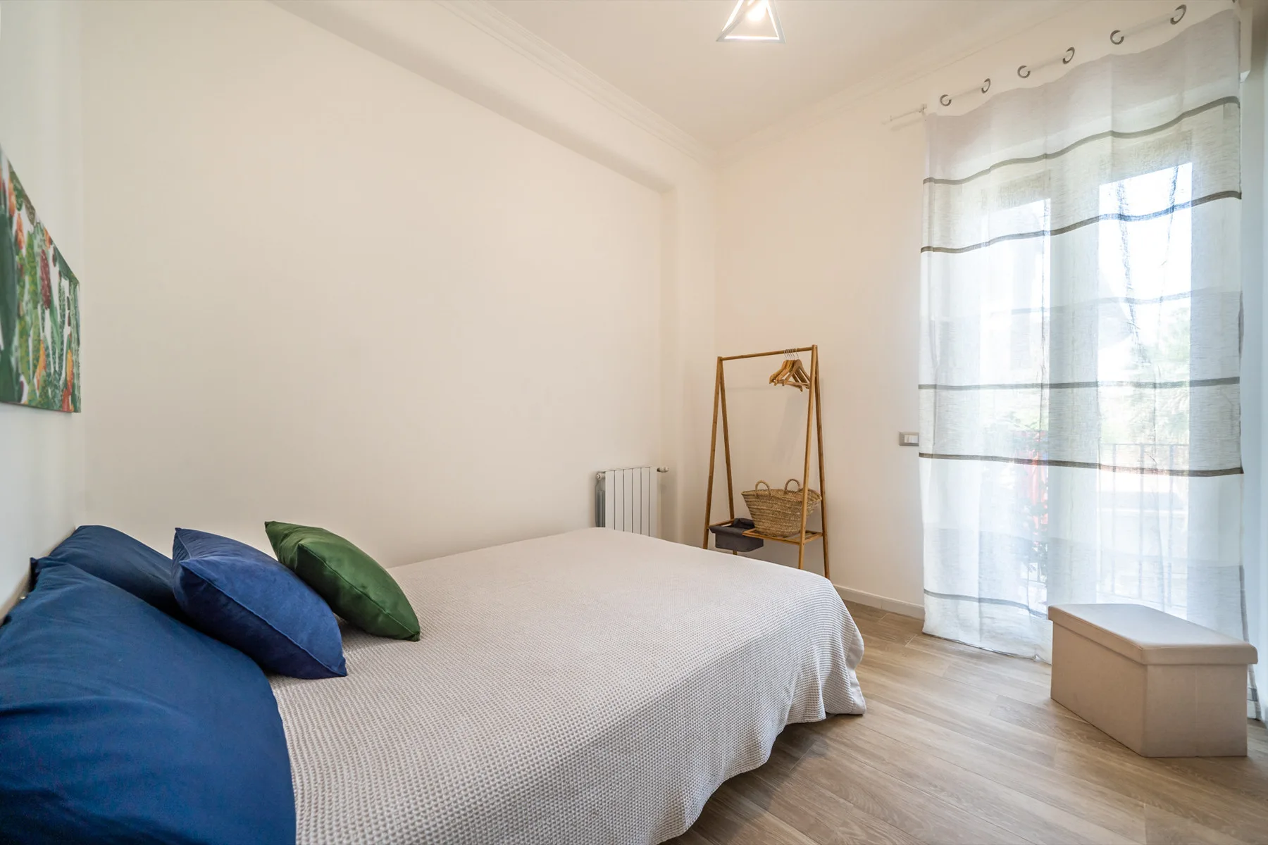 Un dormitorio luminoso con una cama, cojines azules y verdes, y cortinas translúcidas.