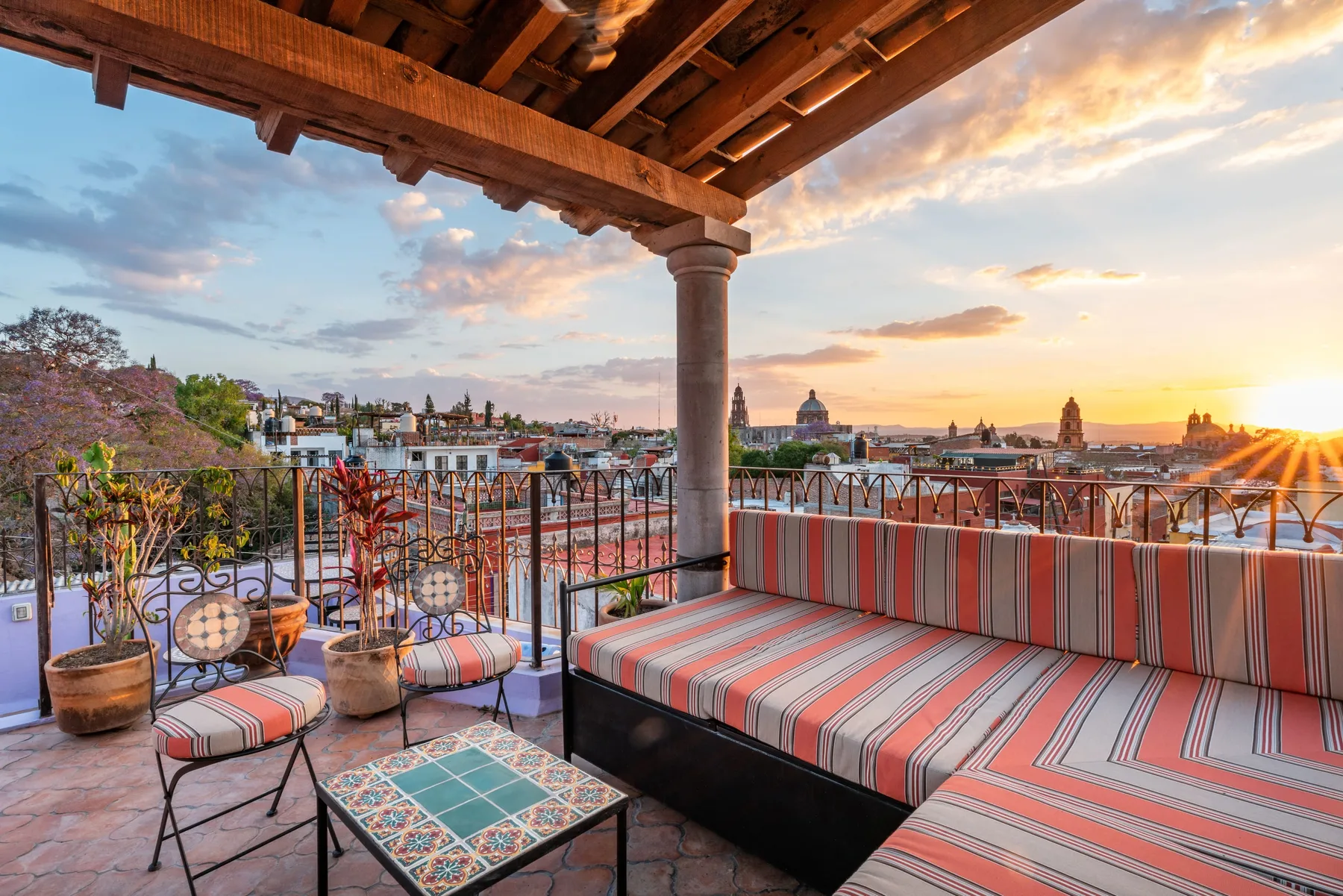 A covered rooftop terrace has striped seating and overlooks a city at sunset.