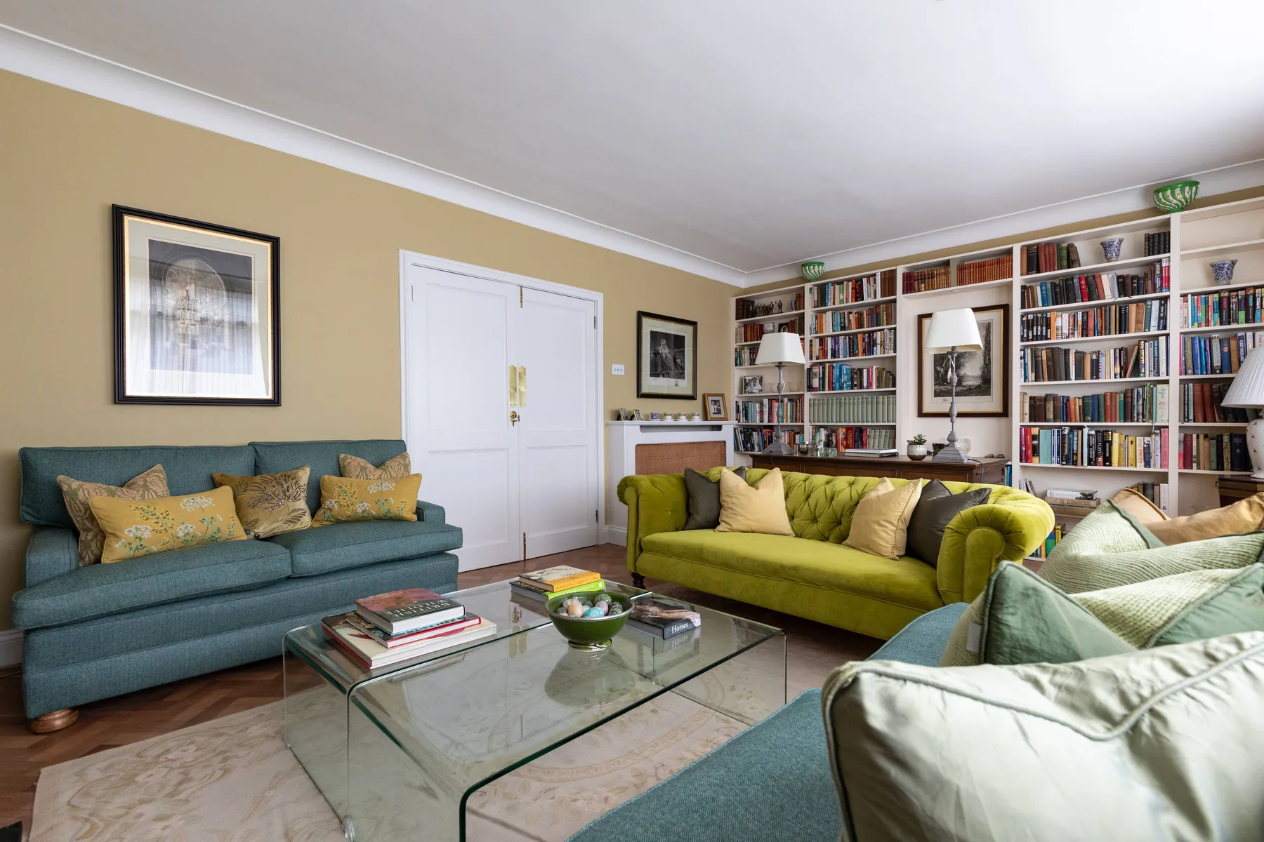A living room with colorful seating, a glass coffee table, and bookshelves lining the walls.