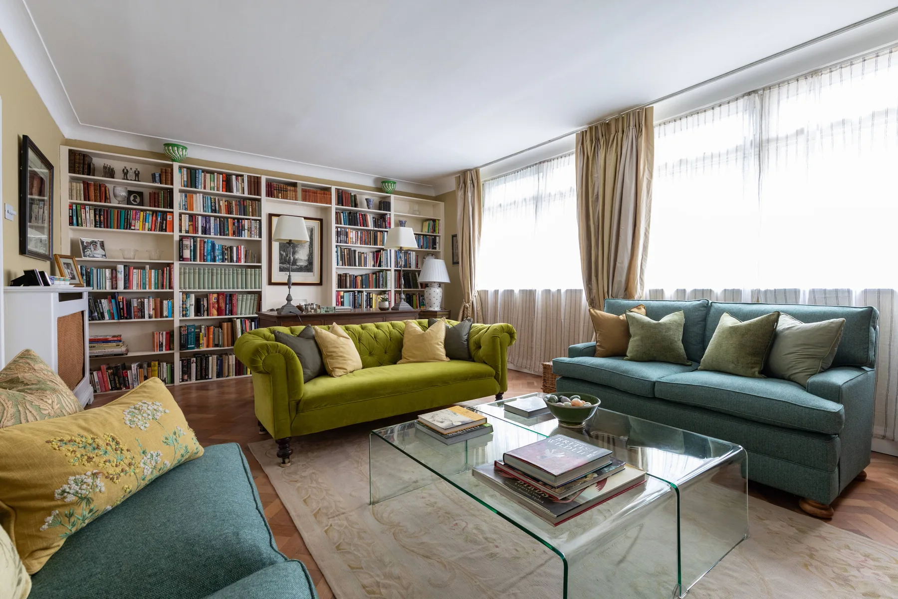 A bright living room with colorful seating, a glass coffee table, and bookshelves.