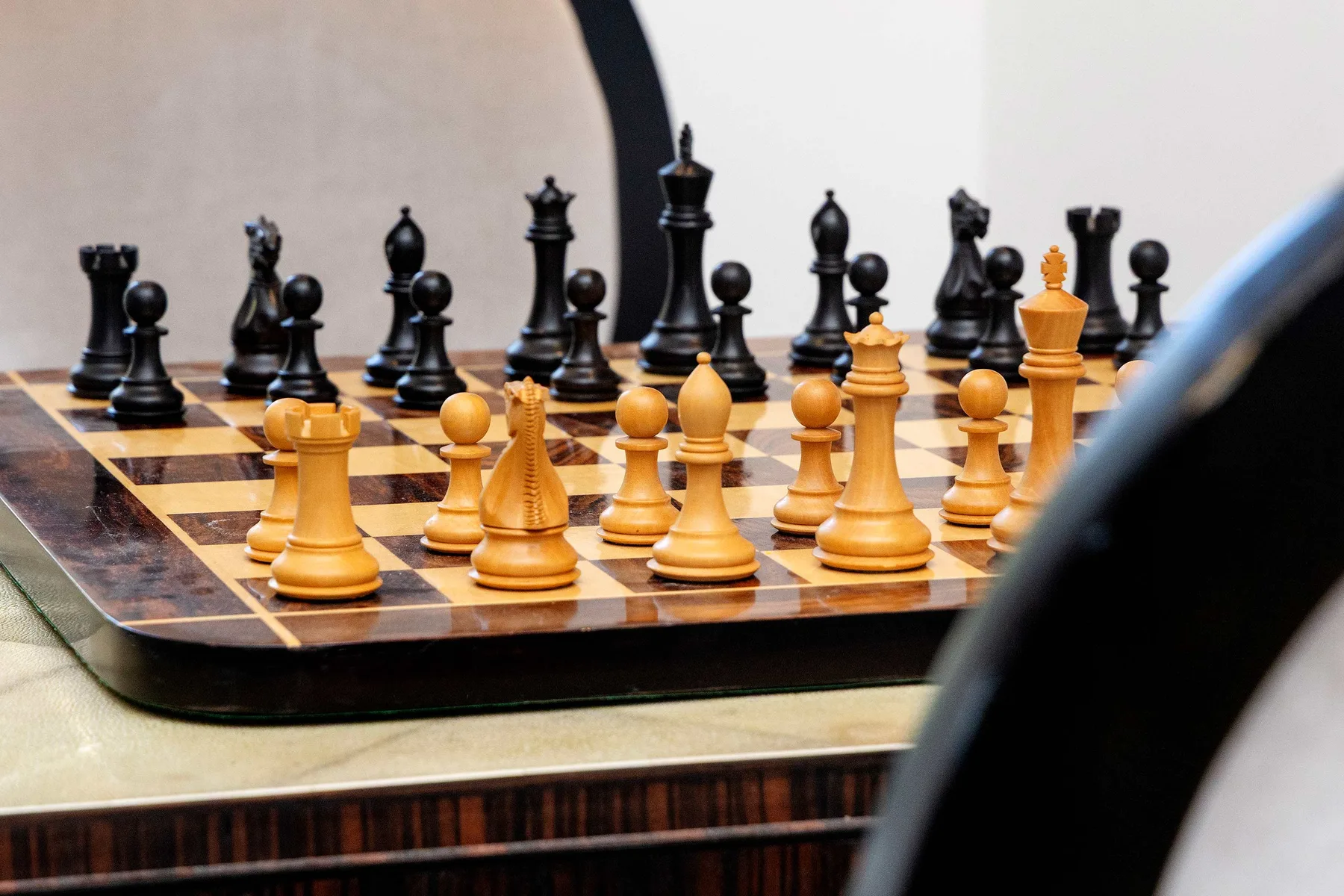 A chessboard with black and tan chess pieces sits on a table.