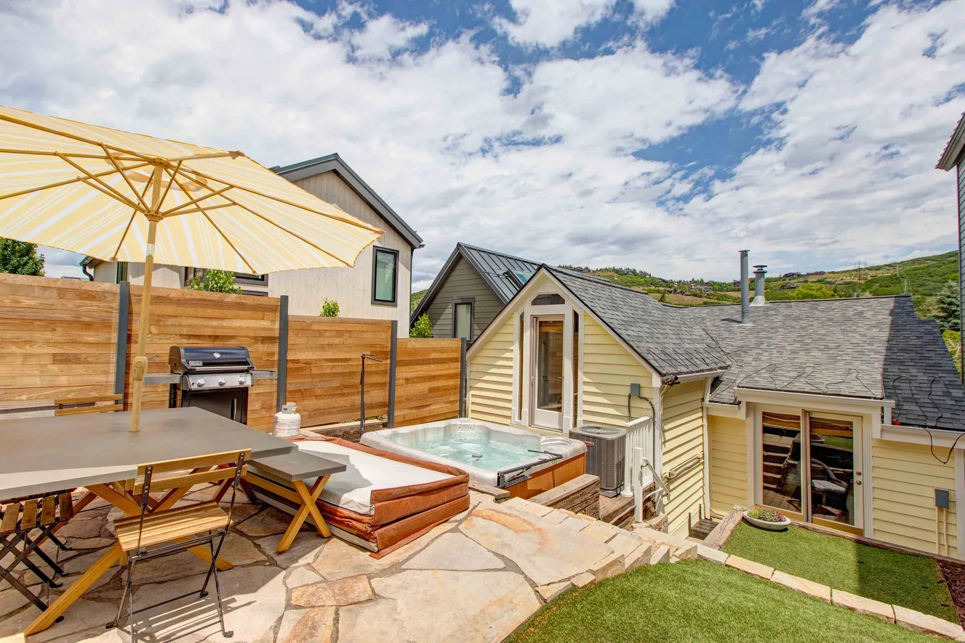 An outdoor patio with a dining table, grill, and hot tub beside a house.