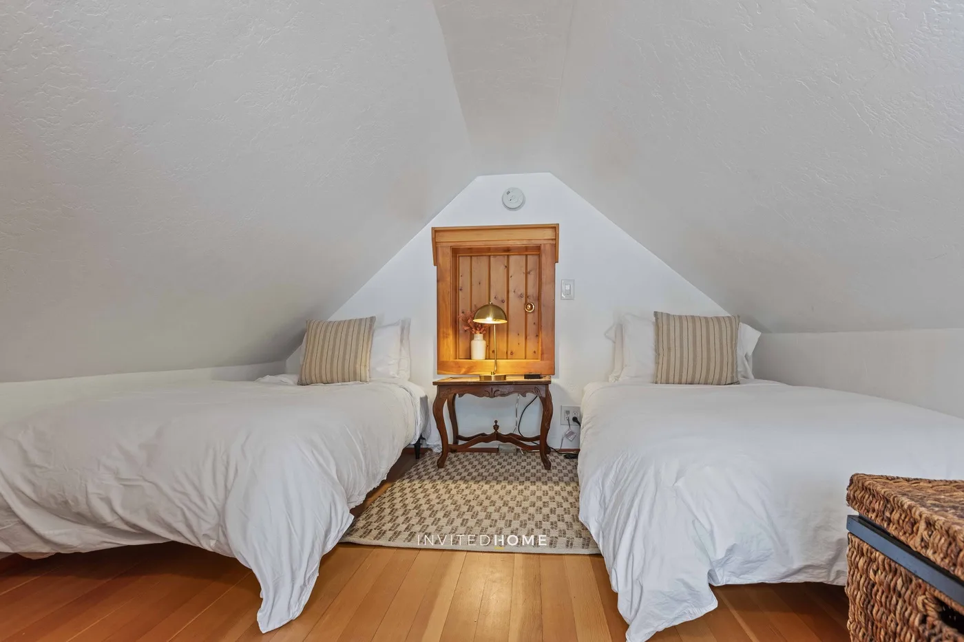 An attic bedroom has two beds beneath sloping ceilings and a wooden window.