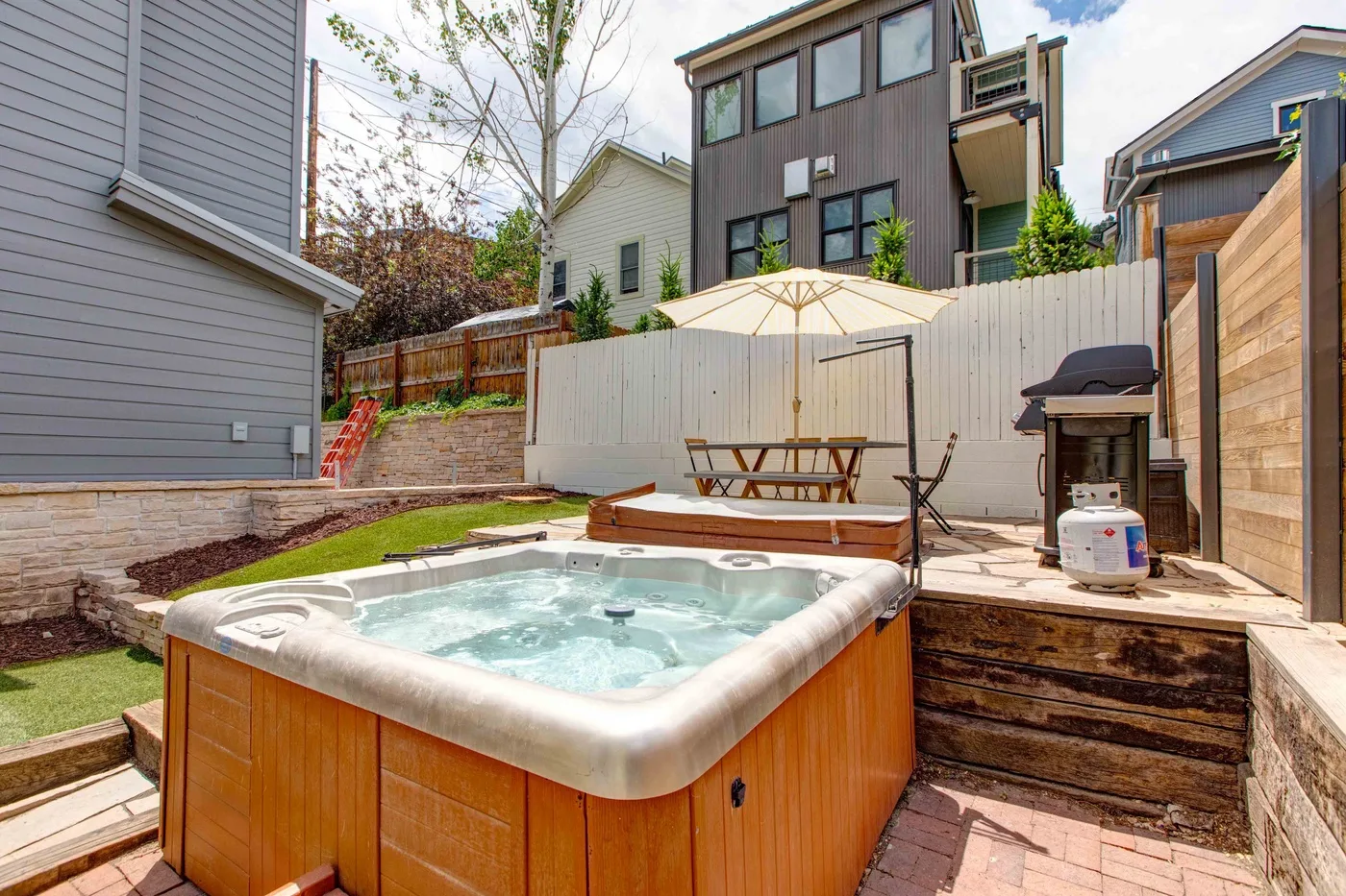 An outdoor hot tub sits beside a small patio with seating and a grill.