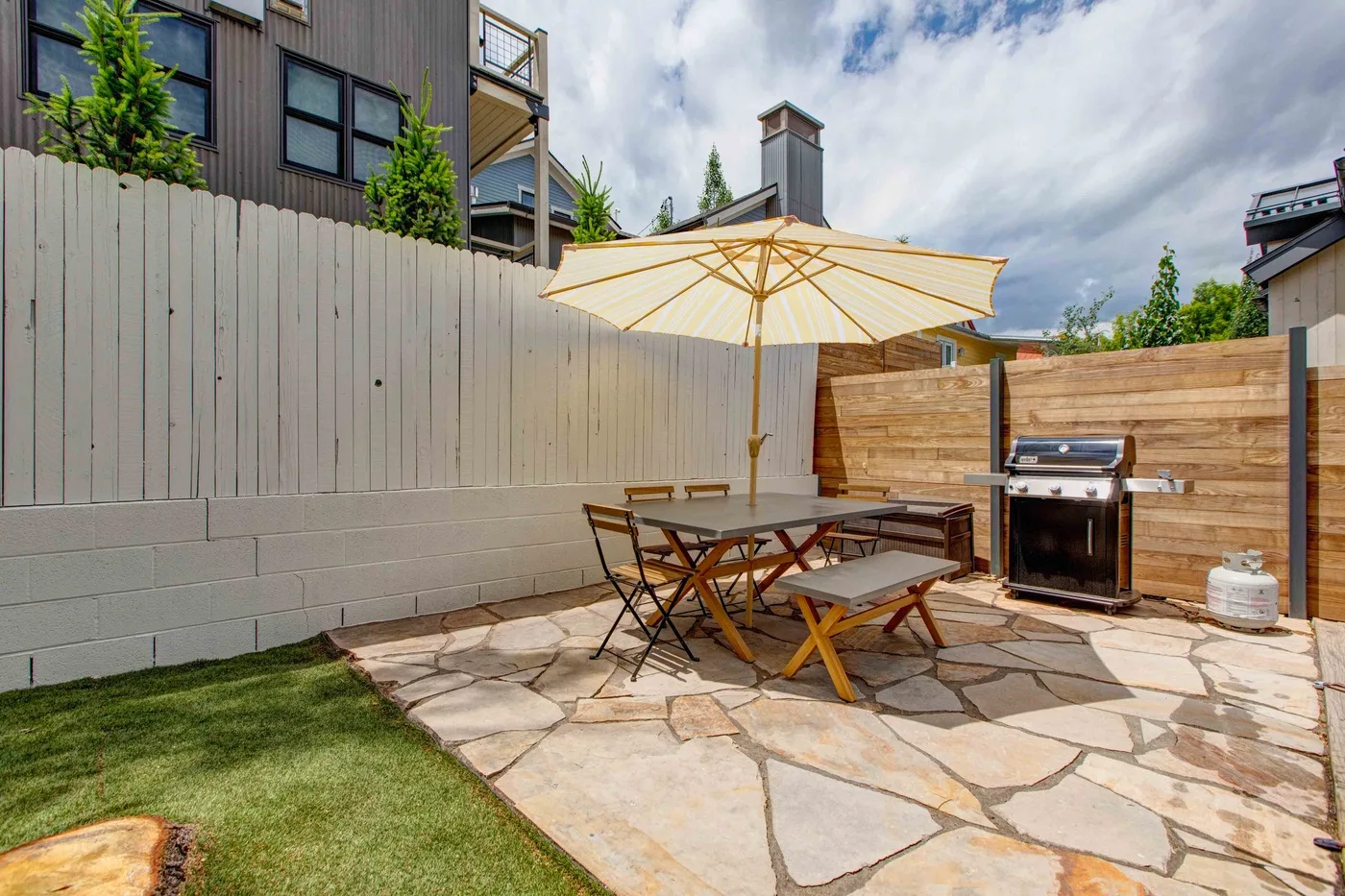 An outdoor patio with a dining table, umbrella, and grill beside a wooden fence.