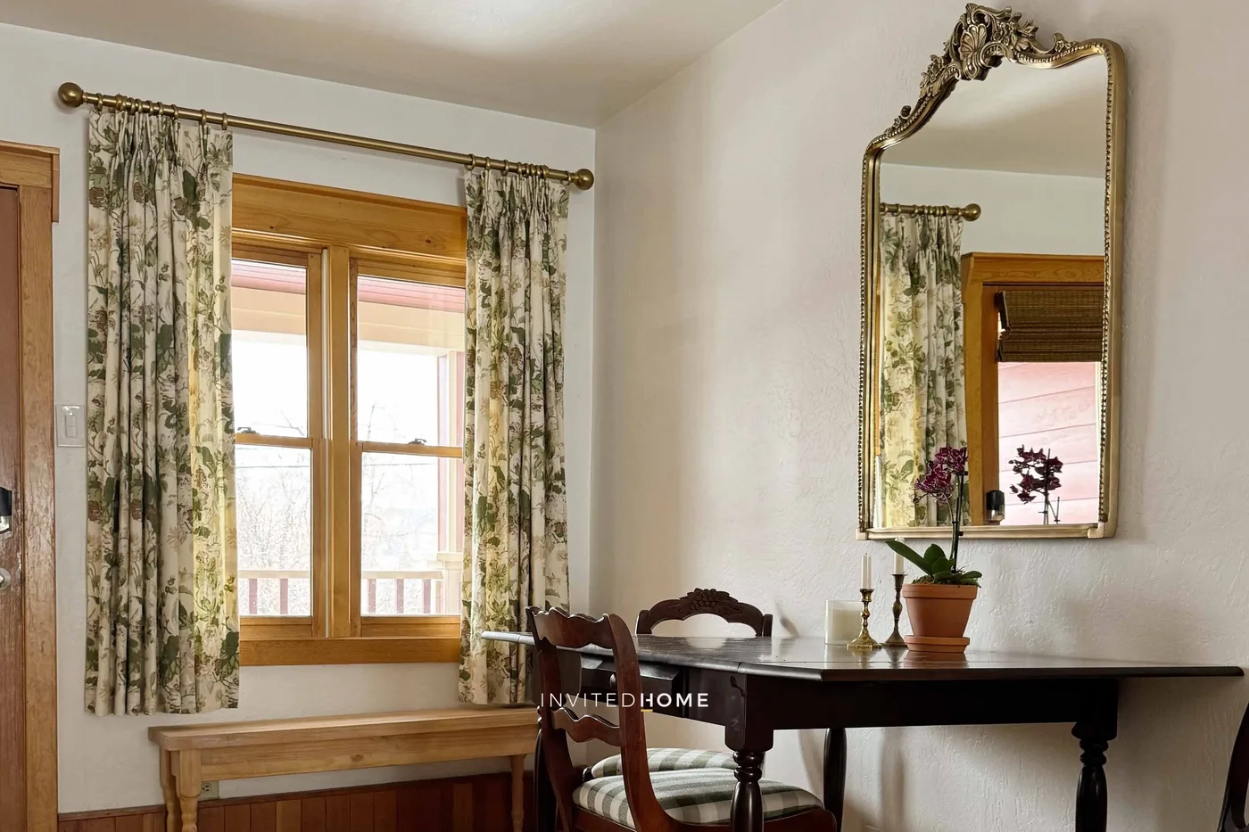 A bedroom corner with a wooden window, patterned curtains, and a dark writing table.