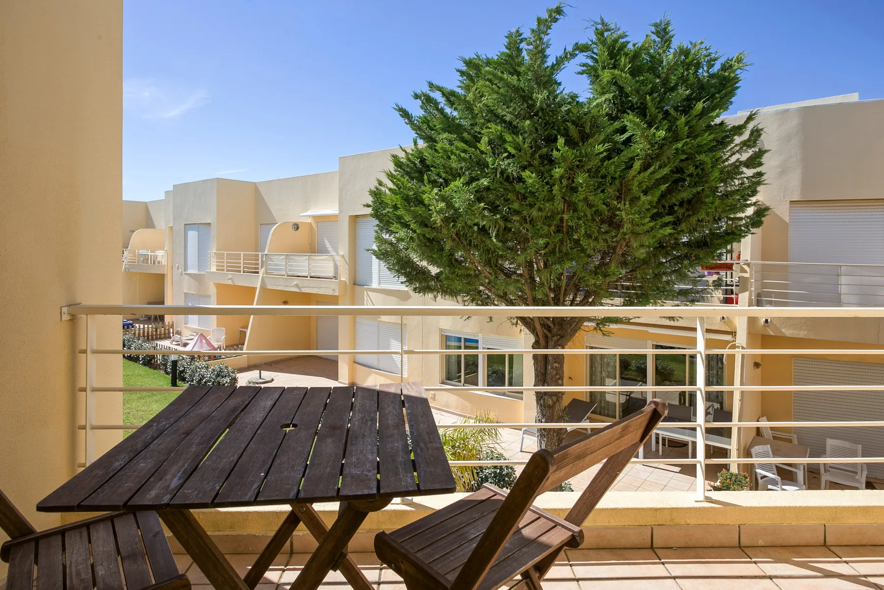 A balcony seating area overlooks neighboring buildings and a leafy tree.