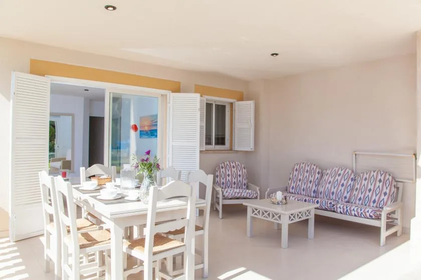 Una luminosa zona de comedor y estar con mesa, asientos y contraventanas abiertas.
