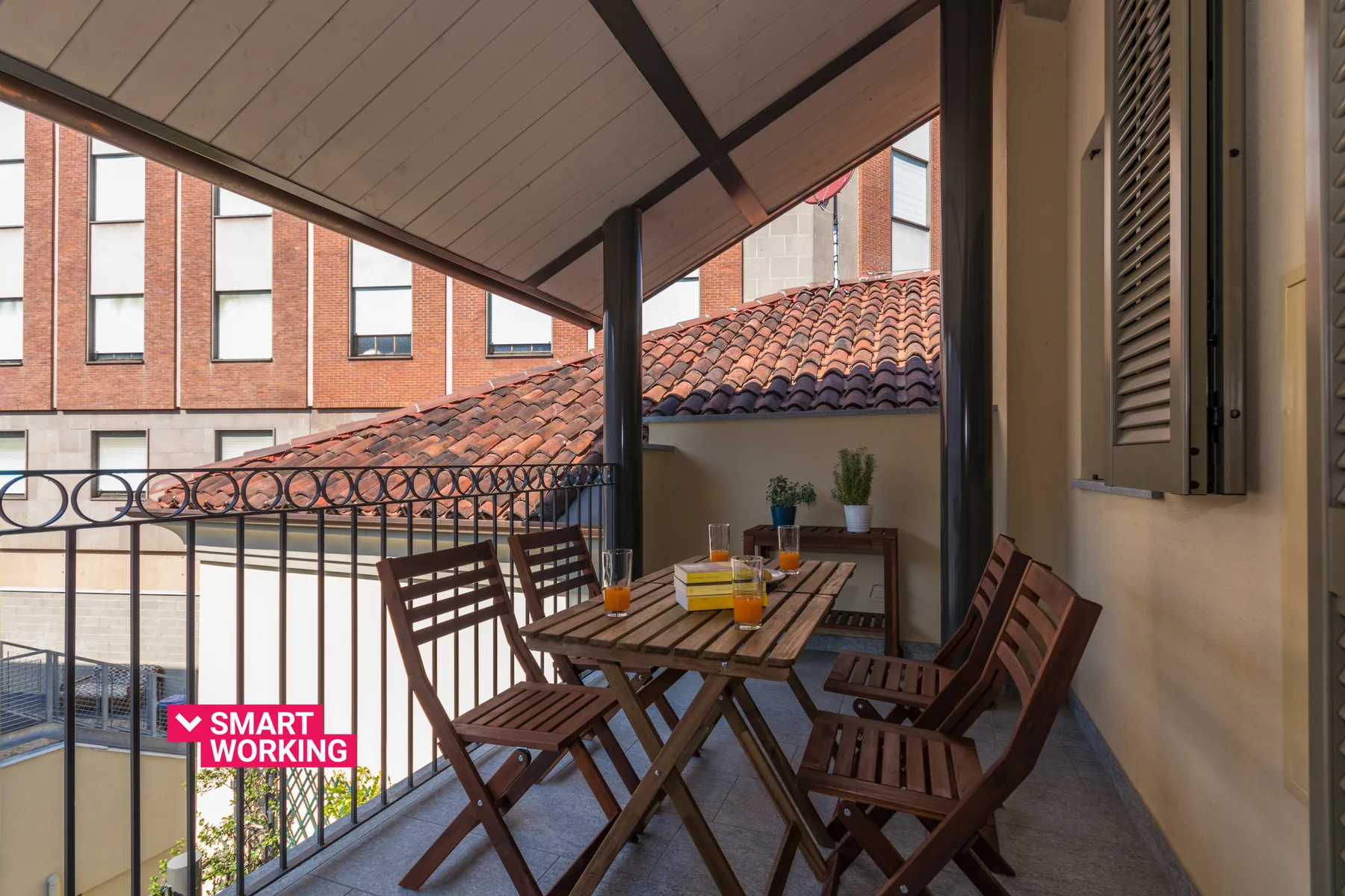 Un balcón cubierto tiene una mesa de comedor y sillas de madera junto a una barandilla metálica.
