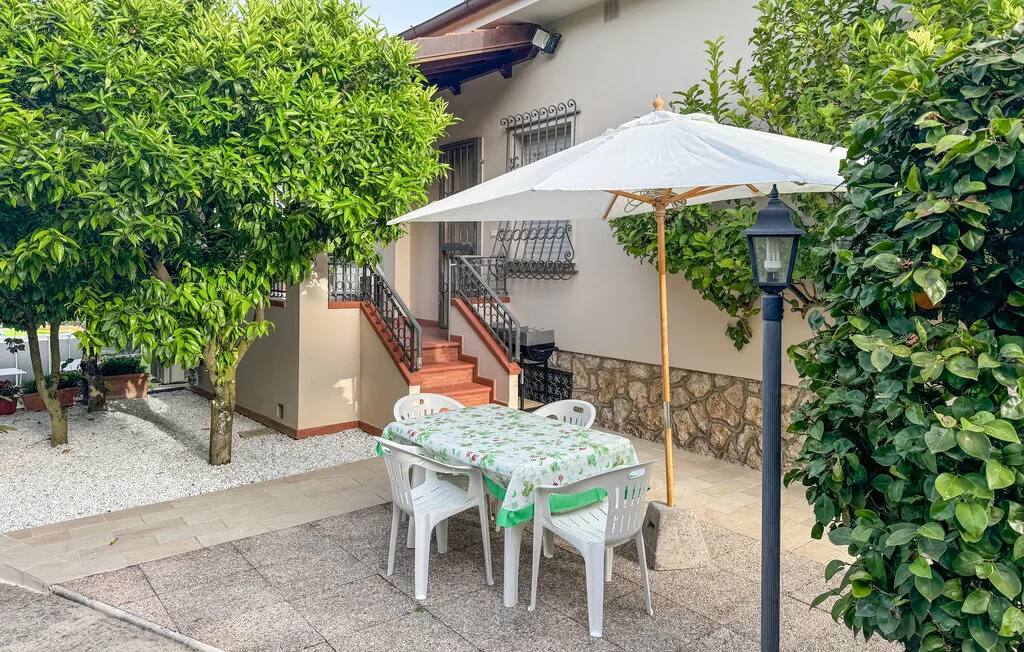Un patio exterior tiene una mesa de comedor bajo una sombrilla blanca, rodeado de vegetación.