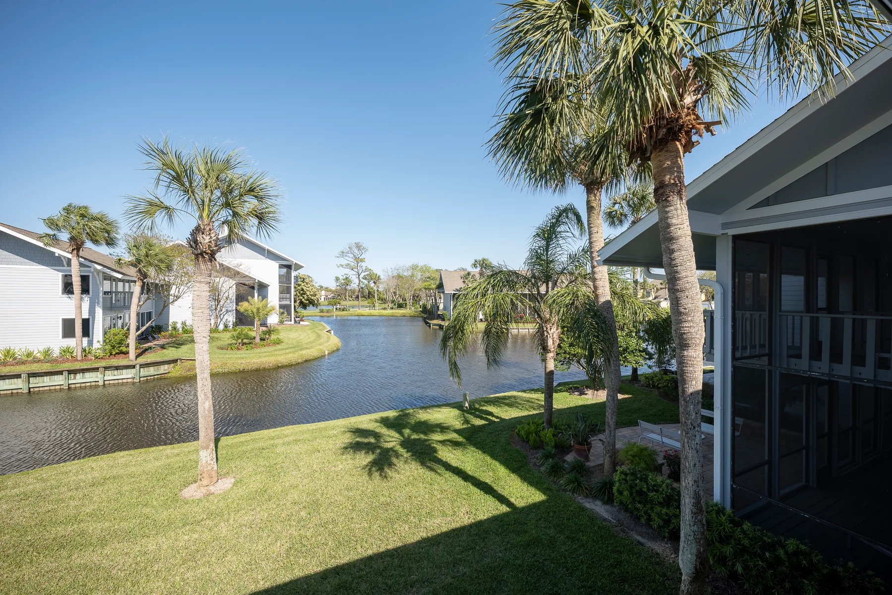 A landscaped waterfront scene with a grassy lawn, palm trees, and buildings beside the water.