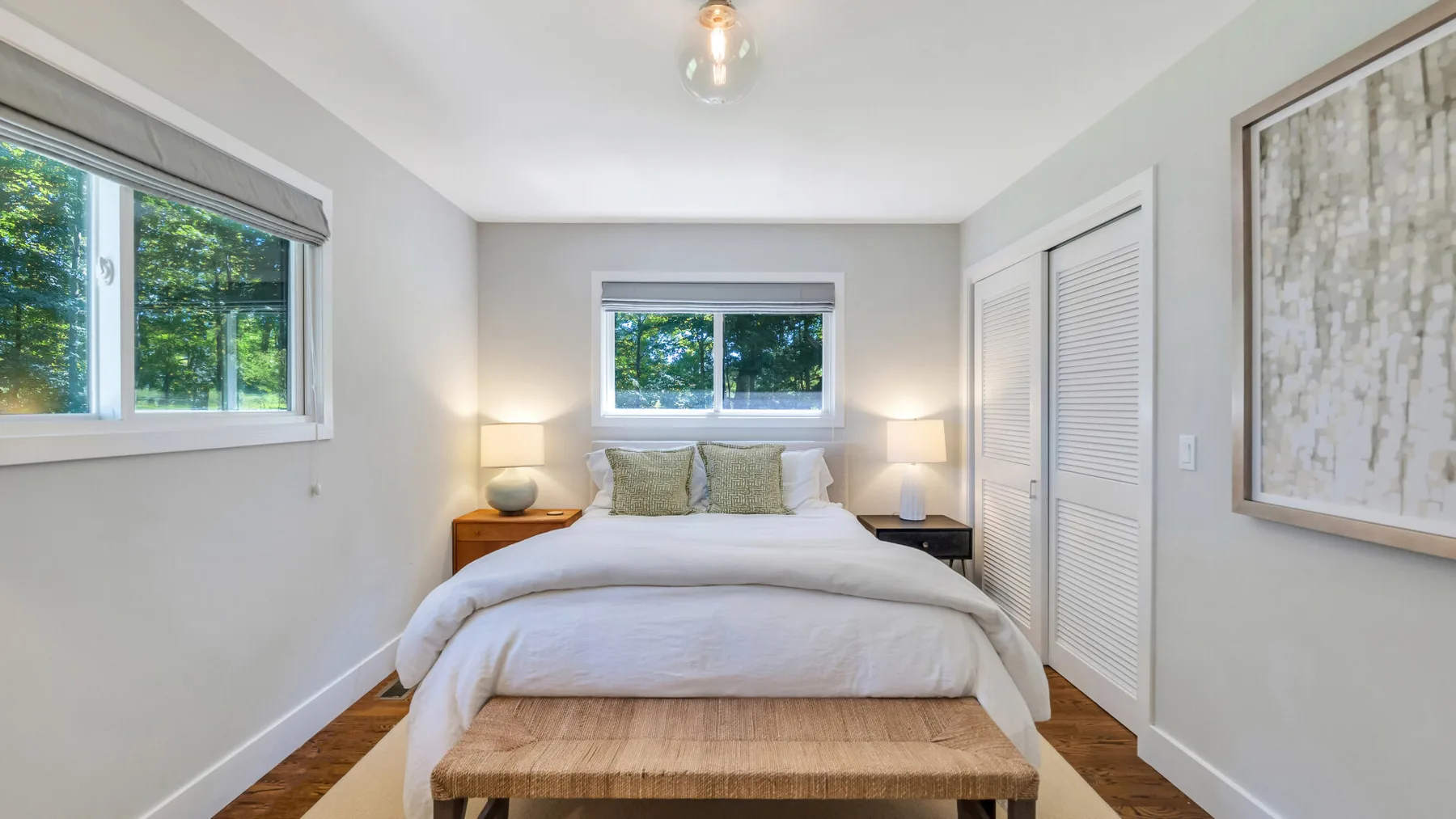 A bright bedroom with a neatly made bed and windows overlooking greenery.