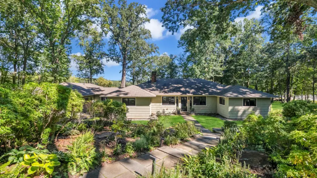 A single-story house sits among trees, with a stone walkway through the garden.