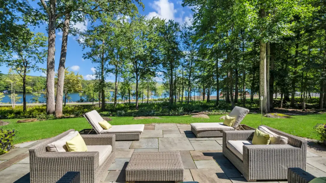 An outdoor patio has woven seating arranged around a low table, with trees and water beyond.