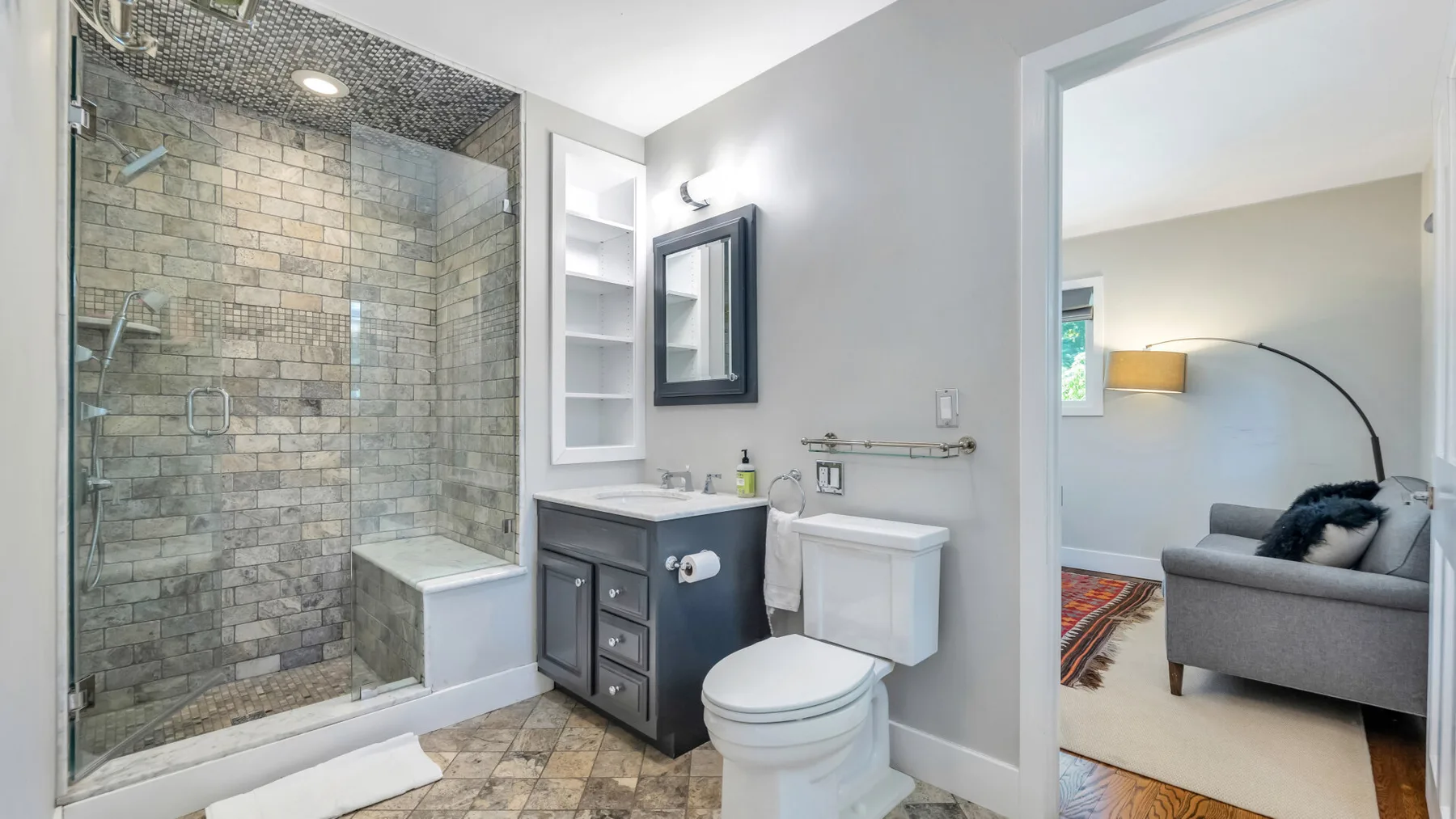 A bathroom with a glass shower, sink, and toilet beside a seating area.