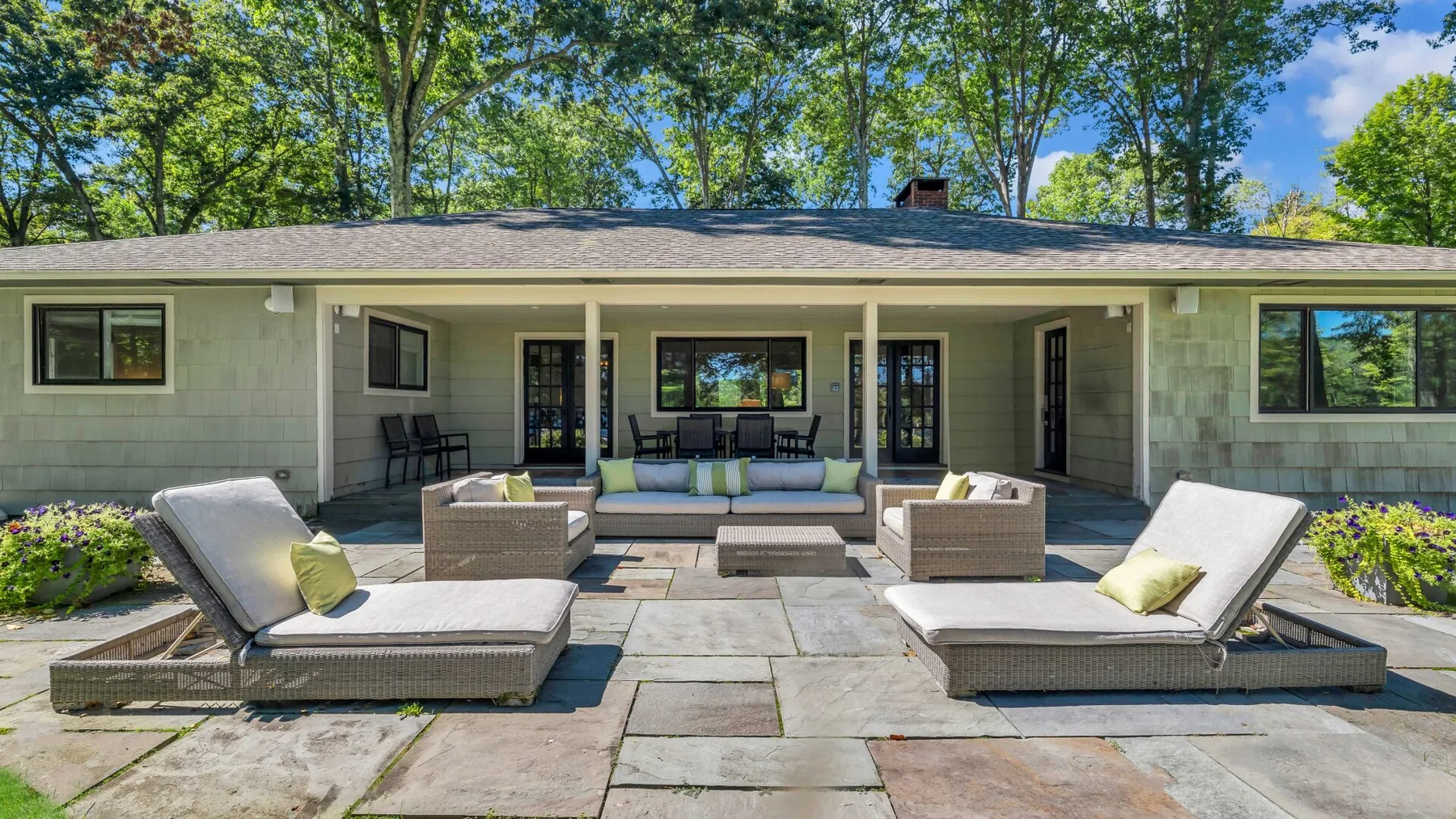 A furnished outdoor patio with lounge seating faces a house and leafy trees.