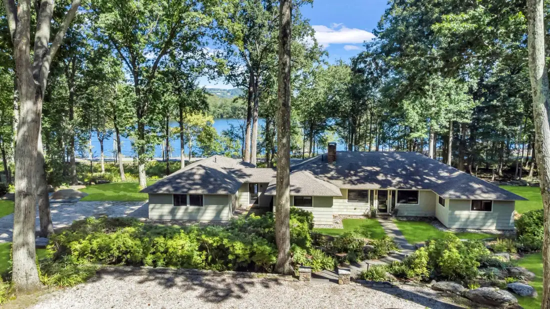 A house sits among trees beside a lake, with a landscaped garden in front.
