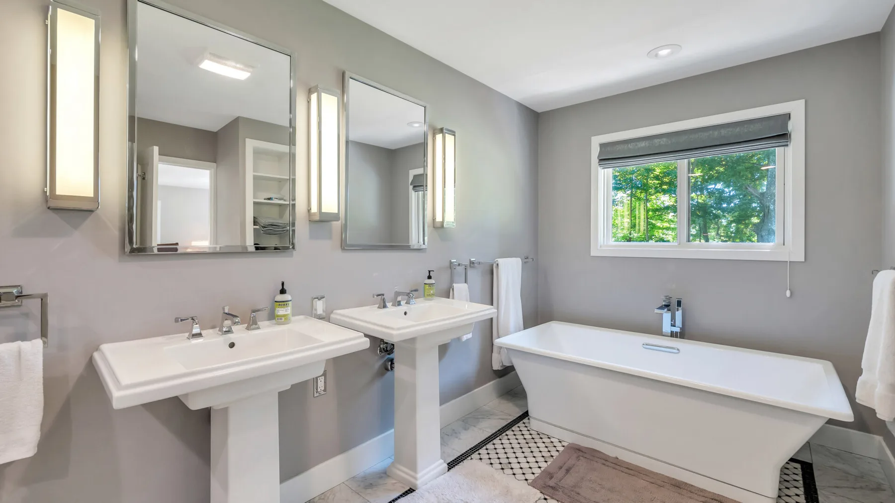 A bright bathroom with two sinks, a bathtub, and a window overlooking greenery.