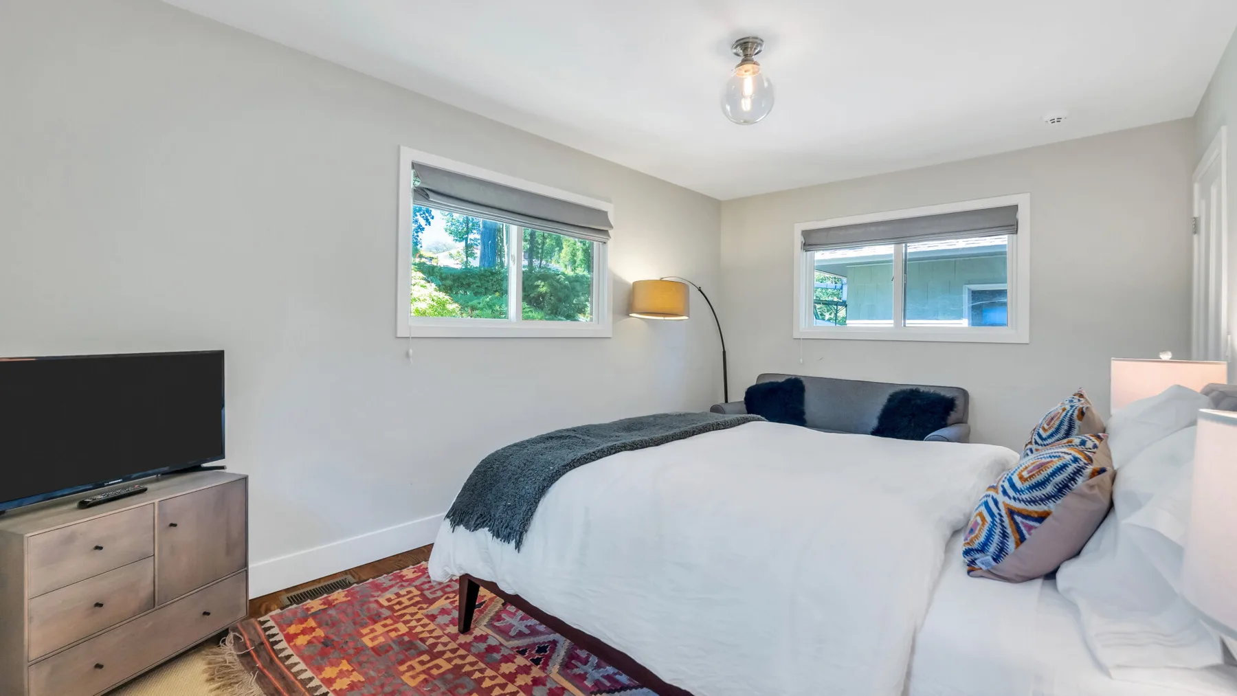 A bright bedroom with a bed, two windows, and a television.