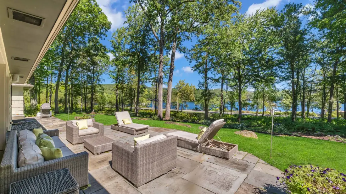 A patio with cushioned seating overlooks a green lawn and a tree-lined lake.