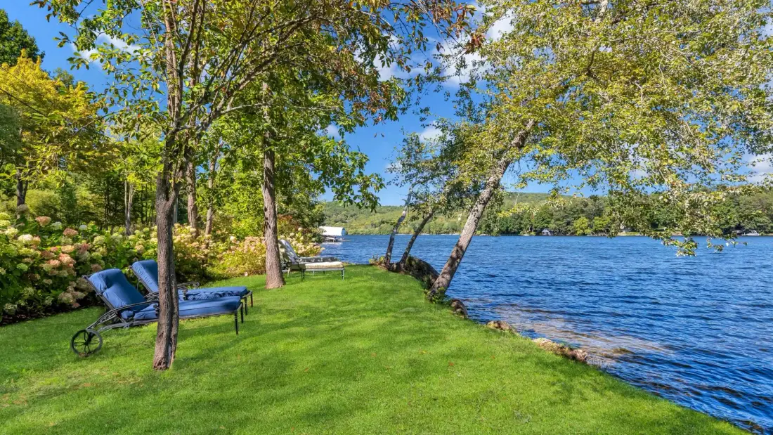 A grassy lakeside lawn is shaded by trees, with seating beside the water.