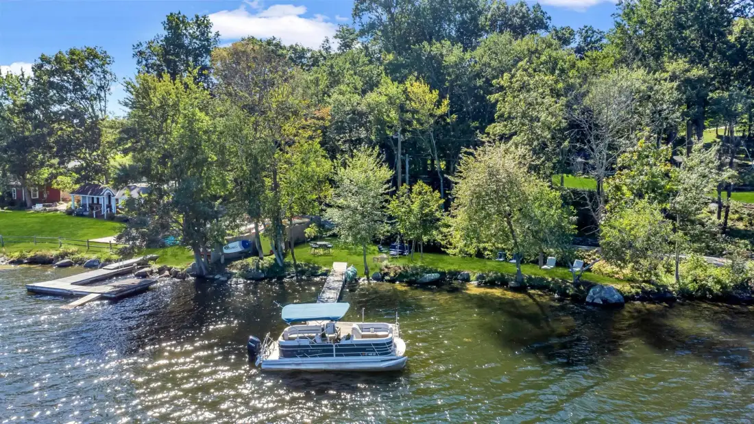 A pontoon boat floats near a wooded shoreline with docks and green lawns.