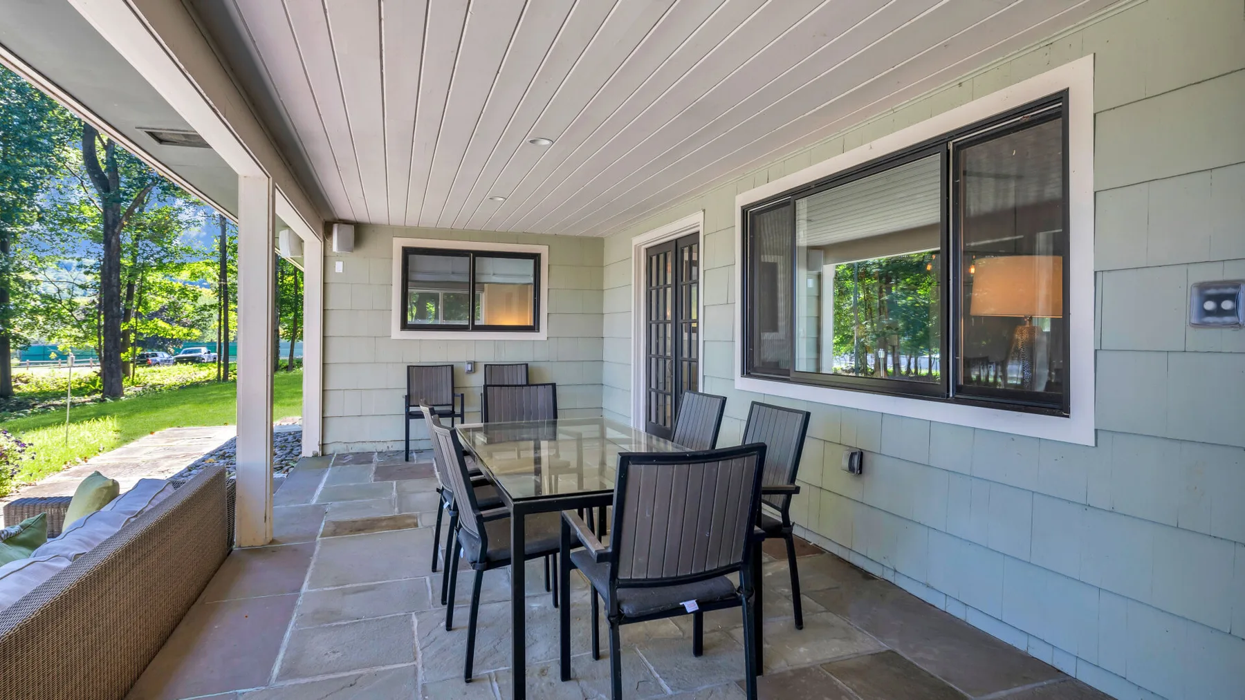 A covered patio has a dining table and chairs beside large windows.