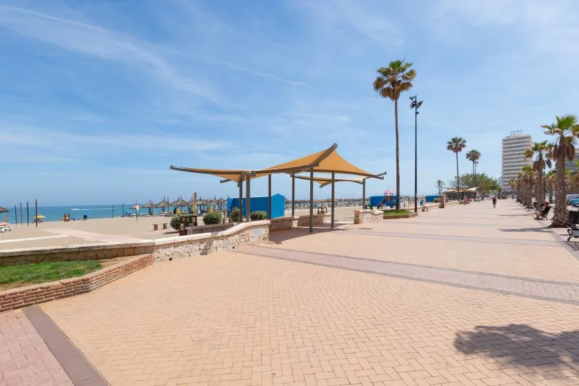 A seaside promenade with a paved walkway, palm trees, and a shade canopy.