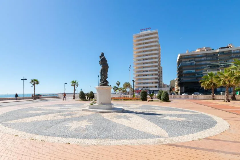 A seaside plaza features a statue, patterned paving, and buildings along the waterfront.
