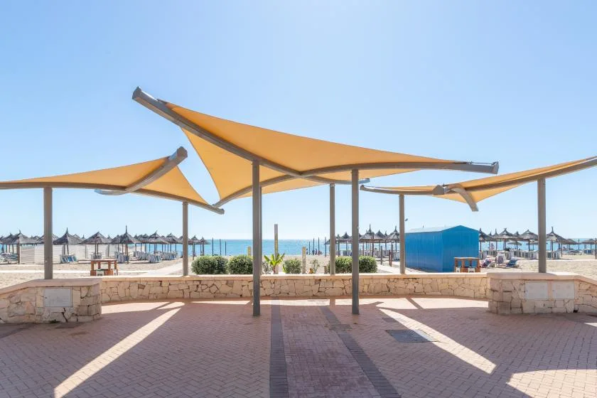 A paved outdoor terrace with orange shade sails overlooks a beach and the sea.