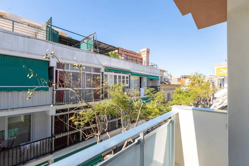 A sunny balcony overlooks neighboring buildings and leafy trees.