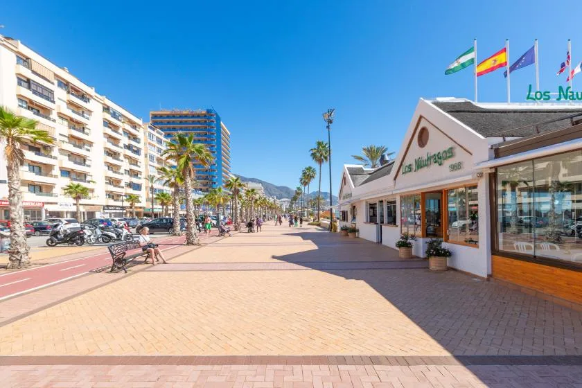 A sunny pedestrian promenade runs past palm trees, shops, and apartment buildings.