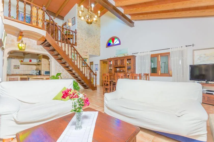 A bright living room with white seating, a wooden staircase, and exposed ceiling beams.