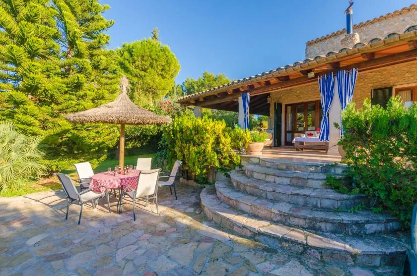 A stone patio beside a house has outdoor seating under a thatched umbrella.