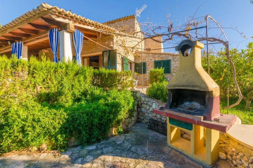 A stone patio with a built-in outdoor grill sits beside a garden and house.
