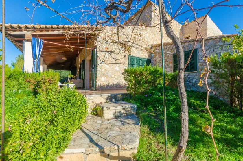 A stone house entrance opens onto a garden path bordered by greenery.