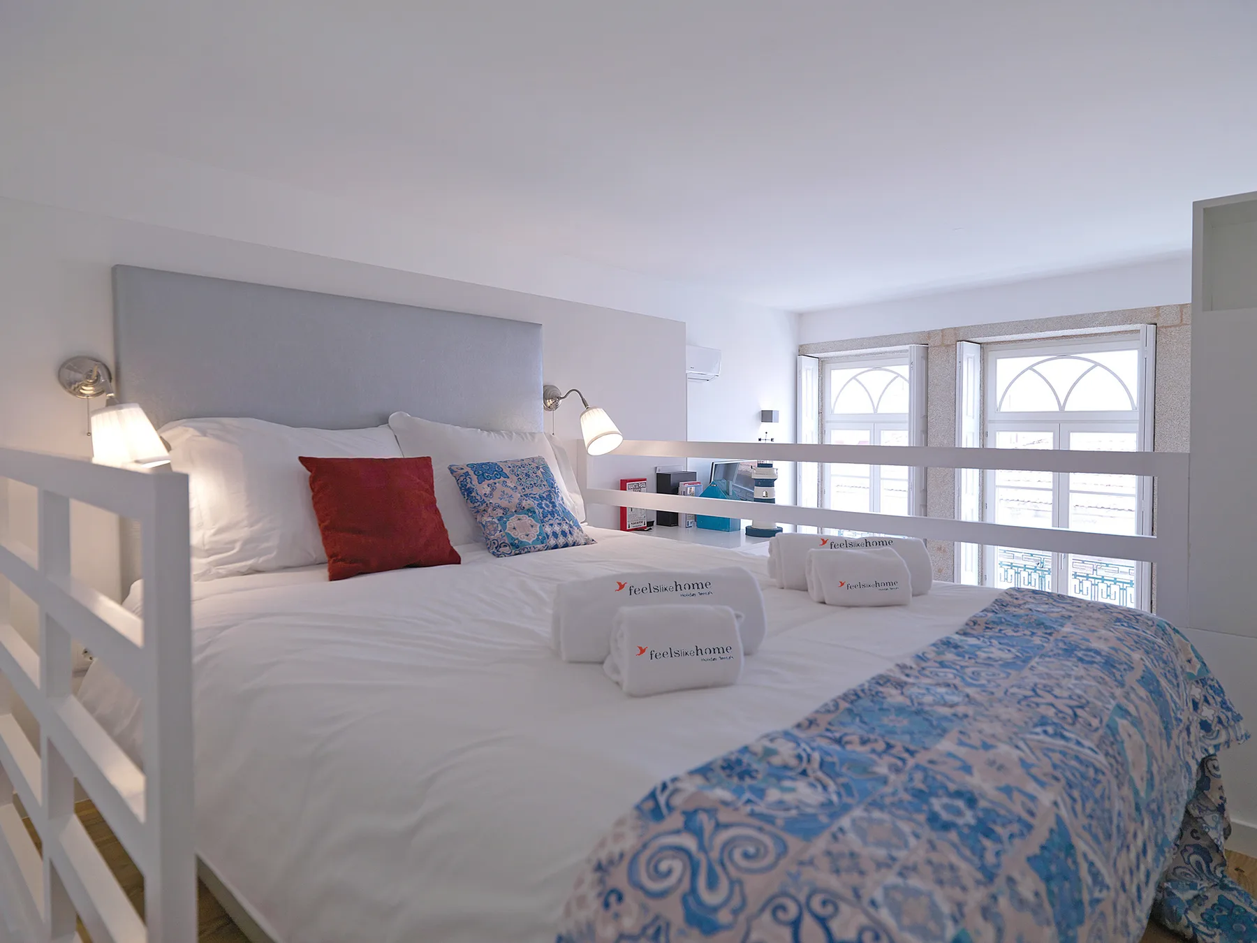 A bright bedroom with white bedding, blue patterned covers, and windows.