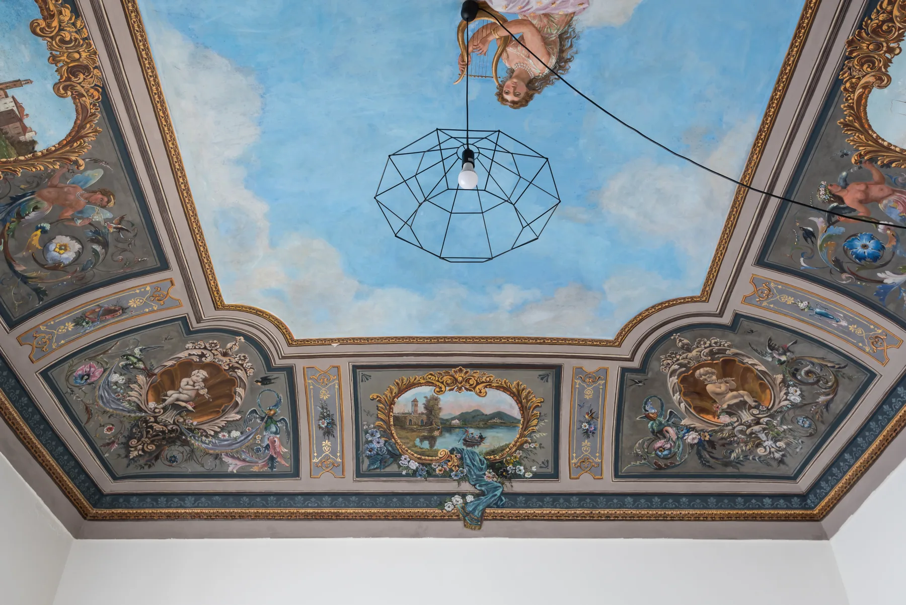 An ornate painted ceiling features a blue sky, framed frescoes, and a geometric pendant light.