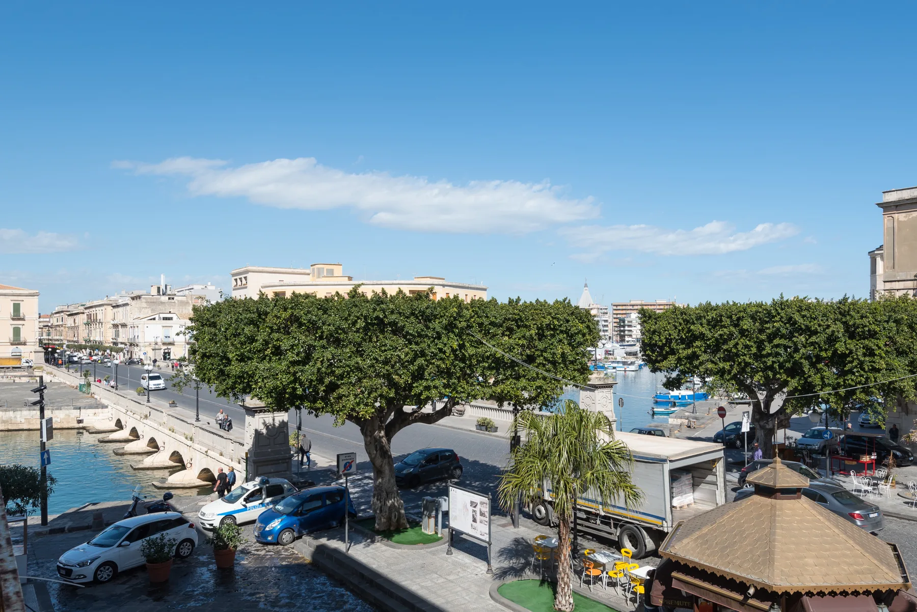 A waterfront street with leafy trees, parked cars, and boats along the harbor.