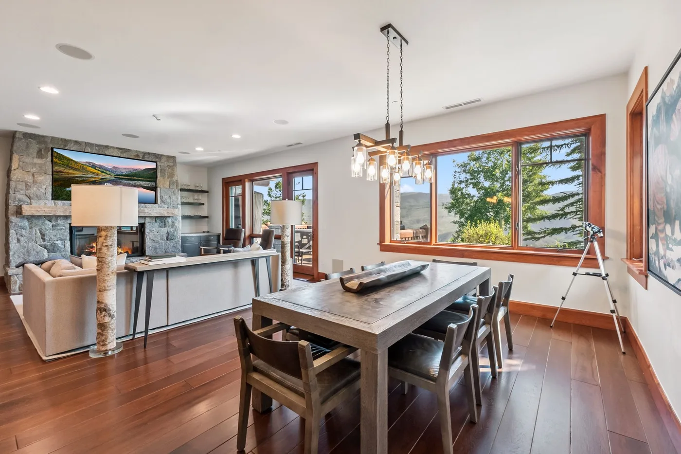 A dining area with a large table beside broad windows overlooking trees.