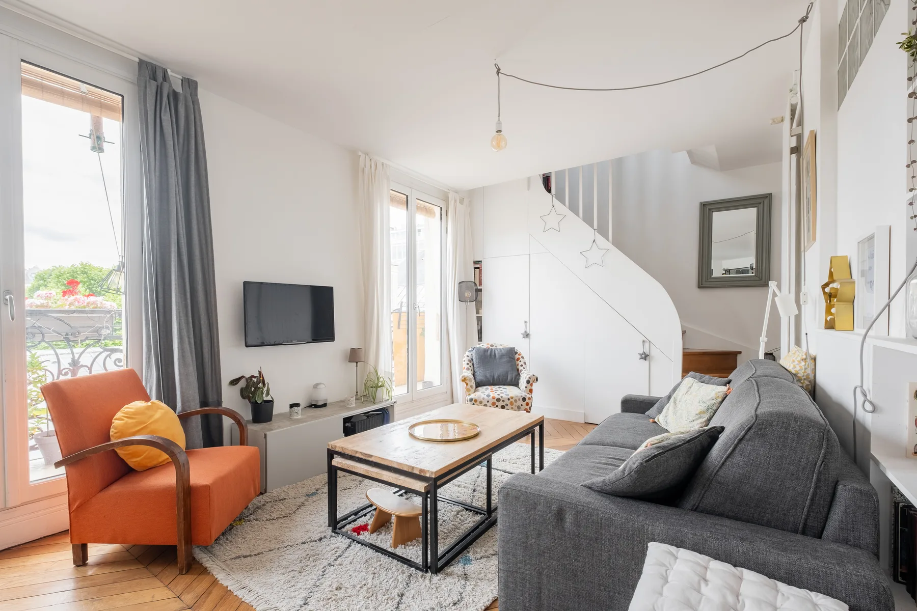 Una sala luminosa con un sofá gris y un sillón naranja junto a las ventanas.