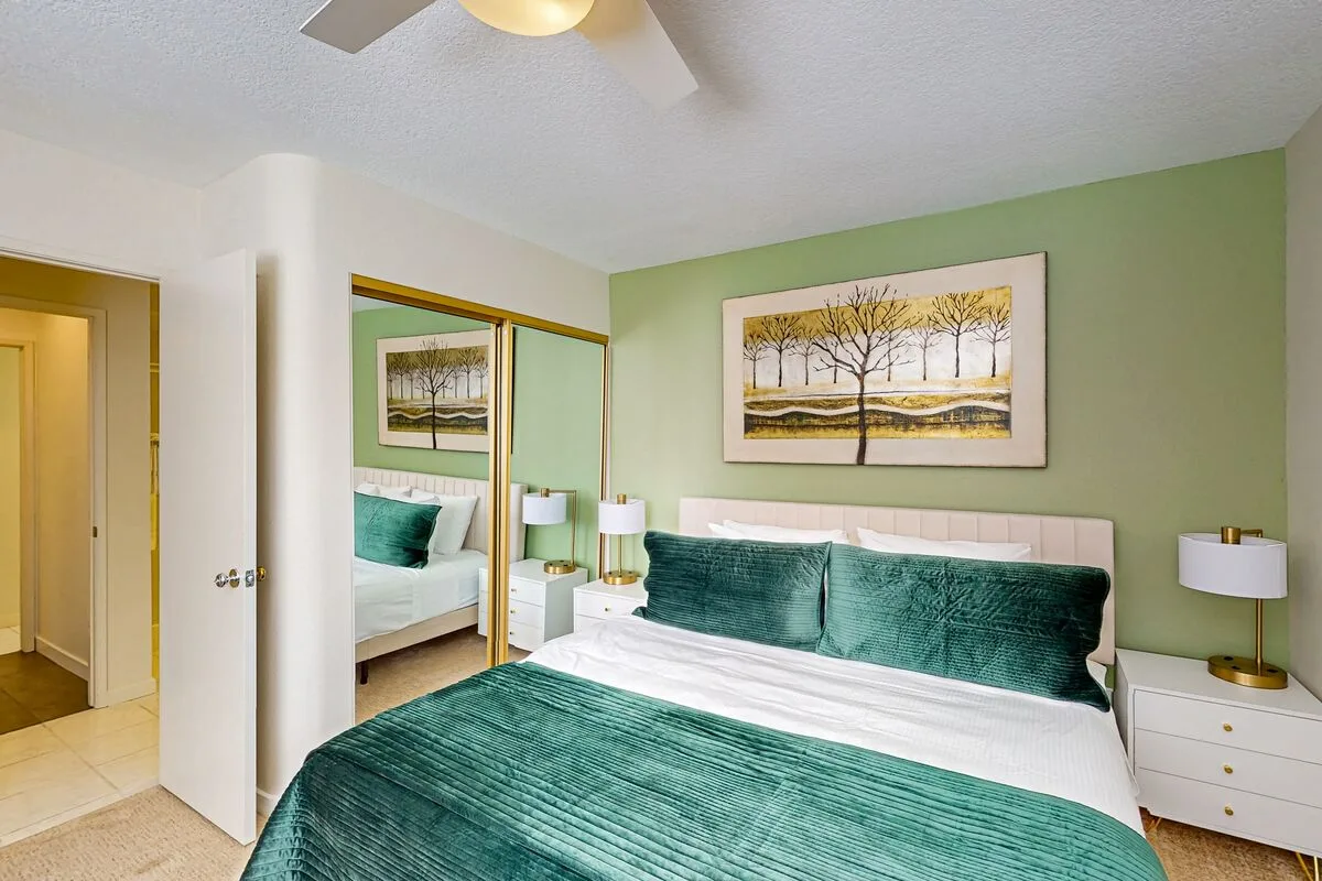 A bedroom with a bed dressed in green bedding and a landscape painting on the wall.