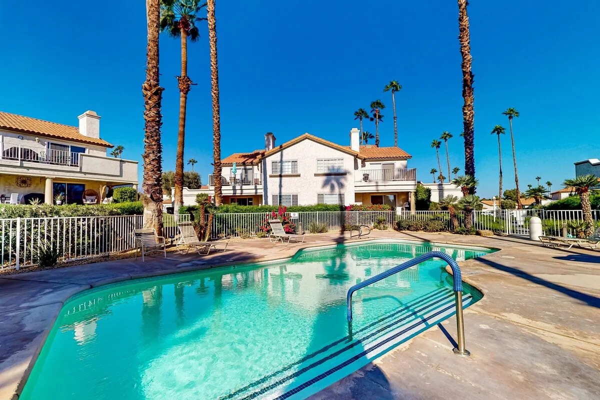 An outdoor swimming pool sits beside a paved deck and low-rise buildings.