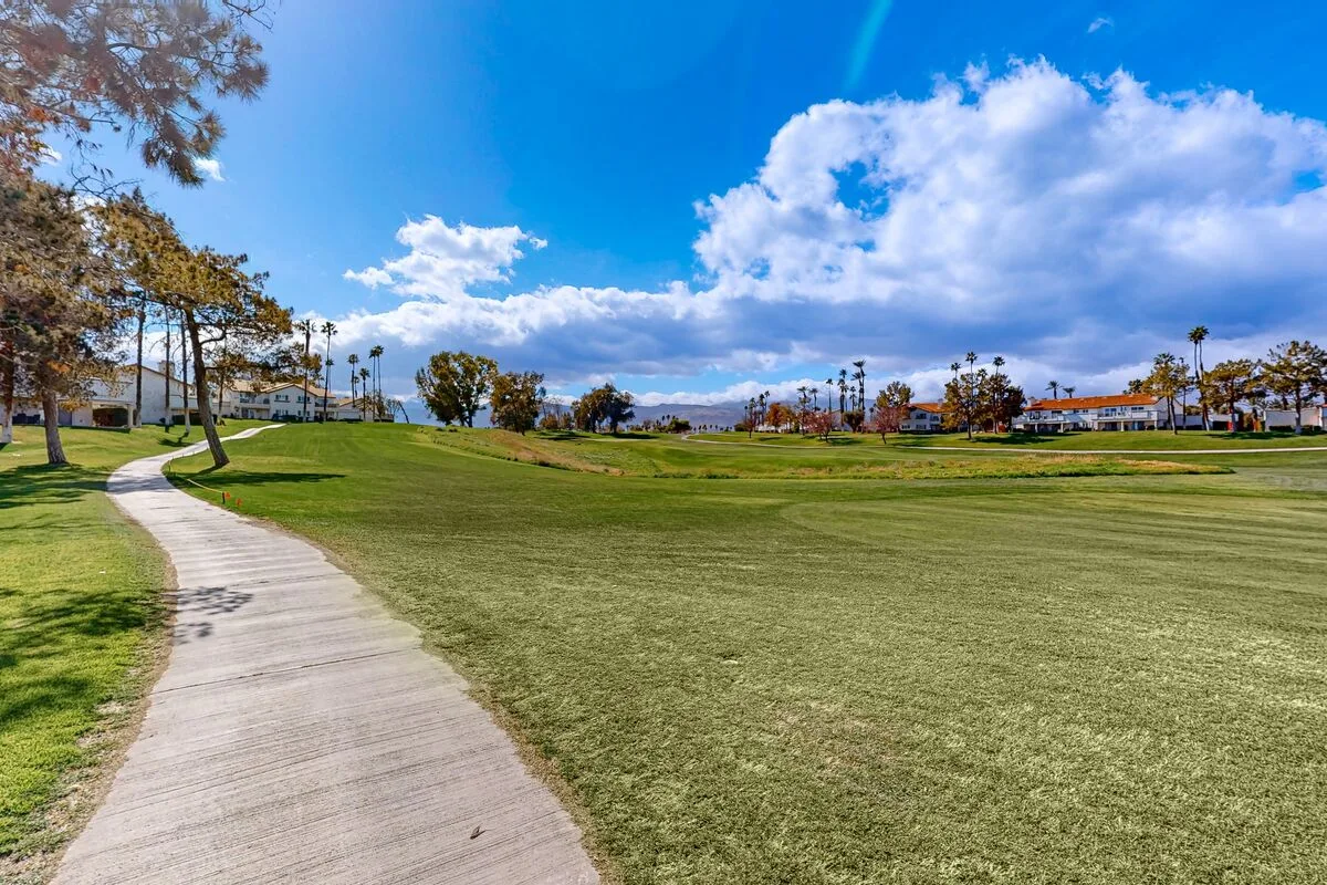 A curving paved path crosses a broad grassy landscape beneath a cloudy blue sky.